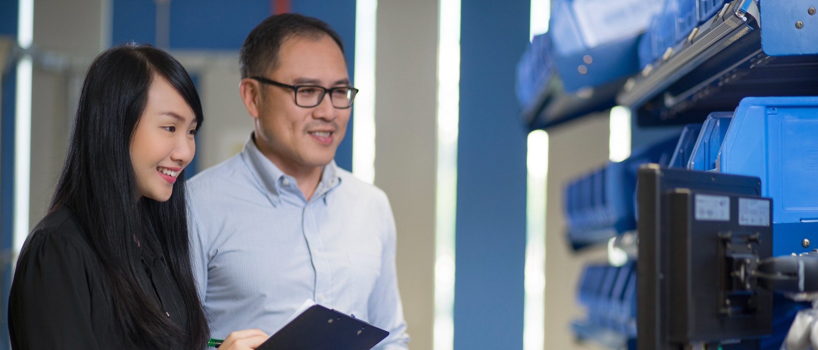 Two people smiling in a warehouse, wearing business attire. One holds a clipboard, while both look at a computer screen